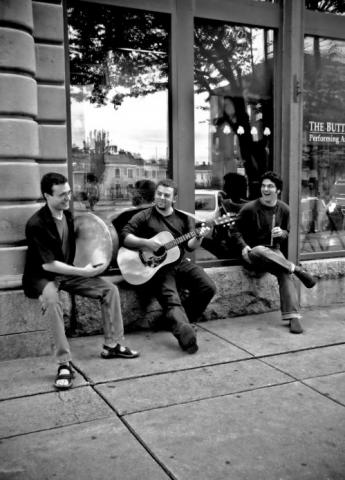 A black and white photo of three of the band members. They are playing a drum, a guitar, and a flute, respectively.