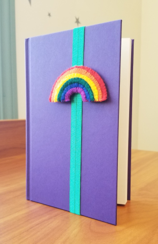 A bookmark made of elastic wrapped around a book cover to hold the reader's place; there is a cartoony felt rainbow attached to the front of the bookmark