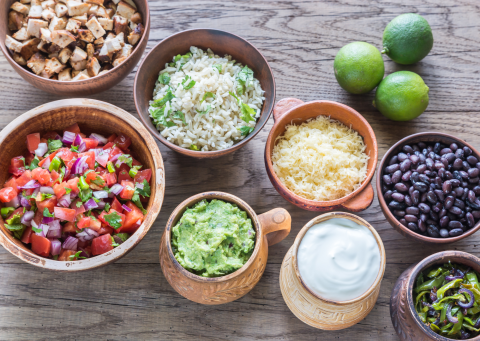 Several bowls of burrito ingredients, including cubed chicken, tomatoes, avocado, cheese, beans, sour cream, and peppers