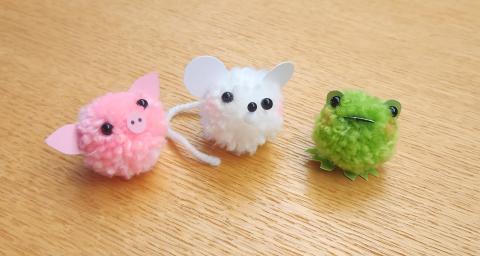 A sample of the craft- a pink pom pom that looks like a pig, a white pom pom that looks like a mouse with a yarn tail, and a green pom pom that looks like a frog.