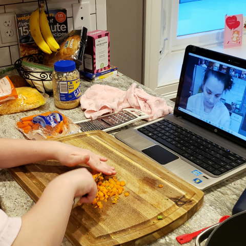 depiction of a tween cooking with the instructor over Zoom; the laptop is in front of the cutting board, and you can see the hands of the tween cutting an orange vegetable