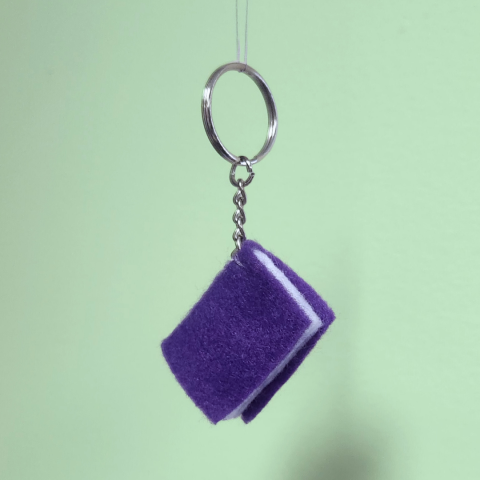 A felt book dangling from a keychain; the book has a purple felt cover and white felt pages.