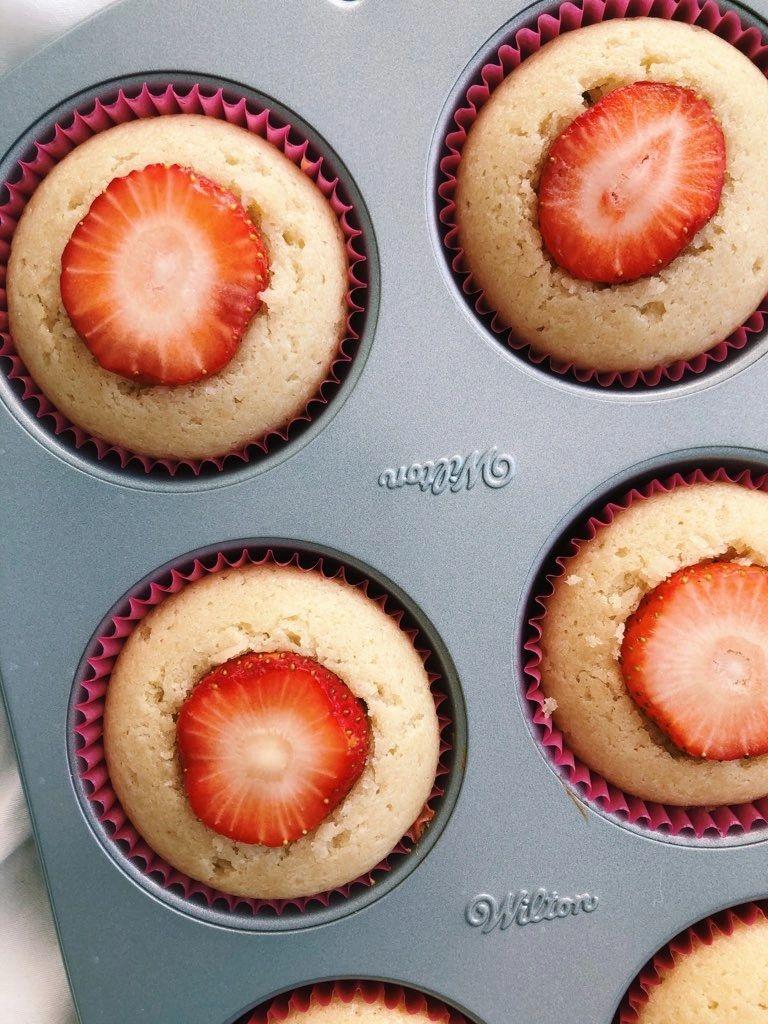 A muffin tin with tan vanilla cupcakes, each cored out to hold a bright red strawberry