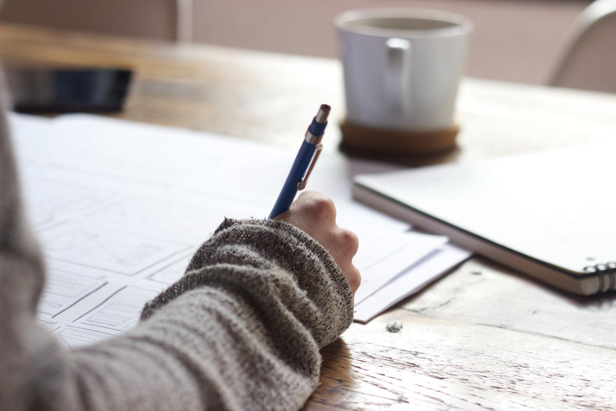 an over the shoulder picture of someone writing an essay with a mug in the background