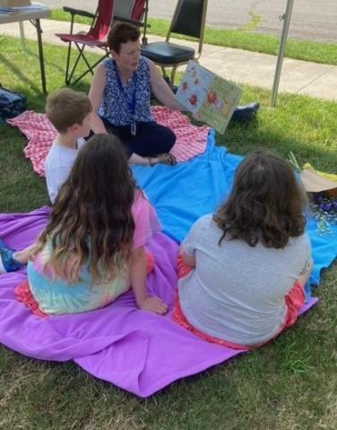 Children's Librarian Jen reading to three children with their backs to the camera; all are seated on picnic blankets on the grass.